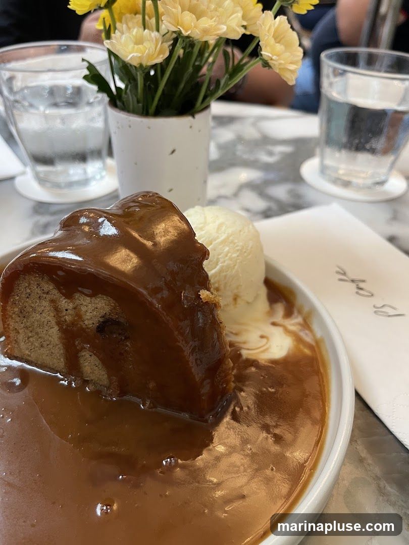 A slice of cake drenched in caramel sauce, served with a scoop of vanilla ice cream. A small vase of yellow flowers and two glasses of water are on the table. A napkin with the text "PS cafe" is visible.