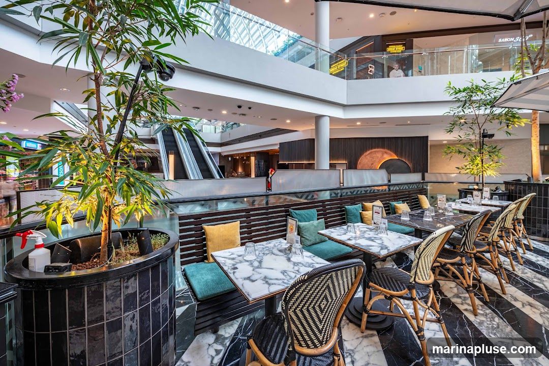 An indoor dining area with marble-topped tables and wicker chairs. The seating features dark wood benches with teal and yellow cushions. A large potted plant with bamboo-like leaves is in the foreground. In the background, escalators and shops are visible on upper levels of a mall.