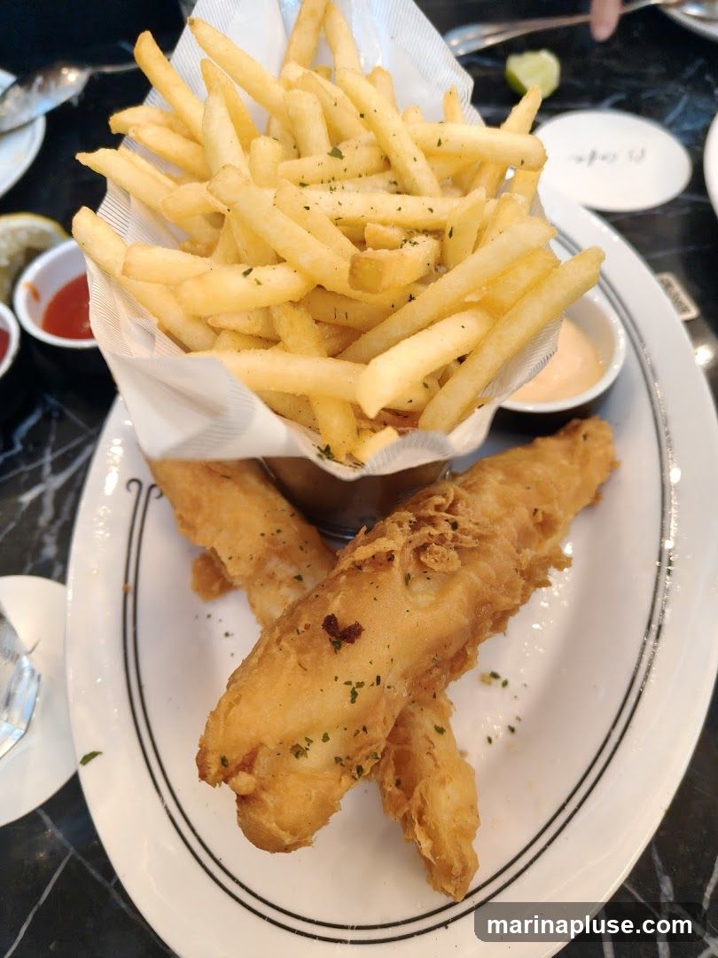 A plate of fish and chips served with french fries and dipping sauces. The fish is golden brown and battered, and the fries are crispy and seasoned with herbs.