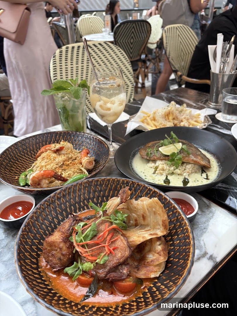 A table at a restaurant is set with several dishes and drinks. In the foreground, a bowl contains a curry dish with meat, flatbread, and tomatoes, served with a side of chili sauce. To the left, a bowl of noodles with shrimp and snap peas is visible. In the background, a plate of french fries and a dish with a piece of fish over rice in a creamy sauce are present. Two drinks, one appearing to be a mojito and another with fruit and ice, are also on the table. The setting appears to be indoors with other diners and chairs visible in the background.