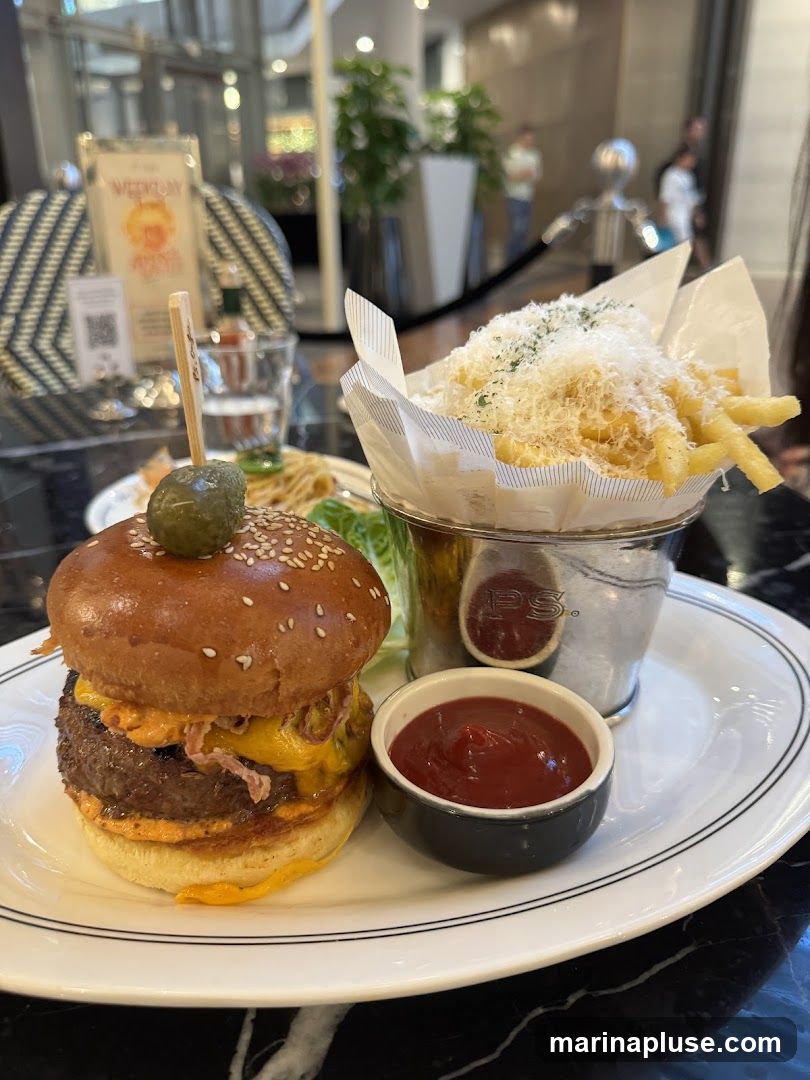 A close-up shot of a gourmet burger and a bucket of fries, served on a white plate with a side of ketchup. The burger is topped with a pickle and sesame seed bun, with melted cheese and sauce. The fries are sprinkled with grated cheese and herbs. In the background, a blurred view of a restaurant interior with plants and patrons can be seen.