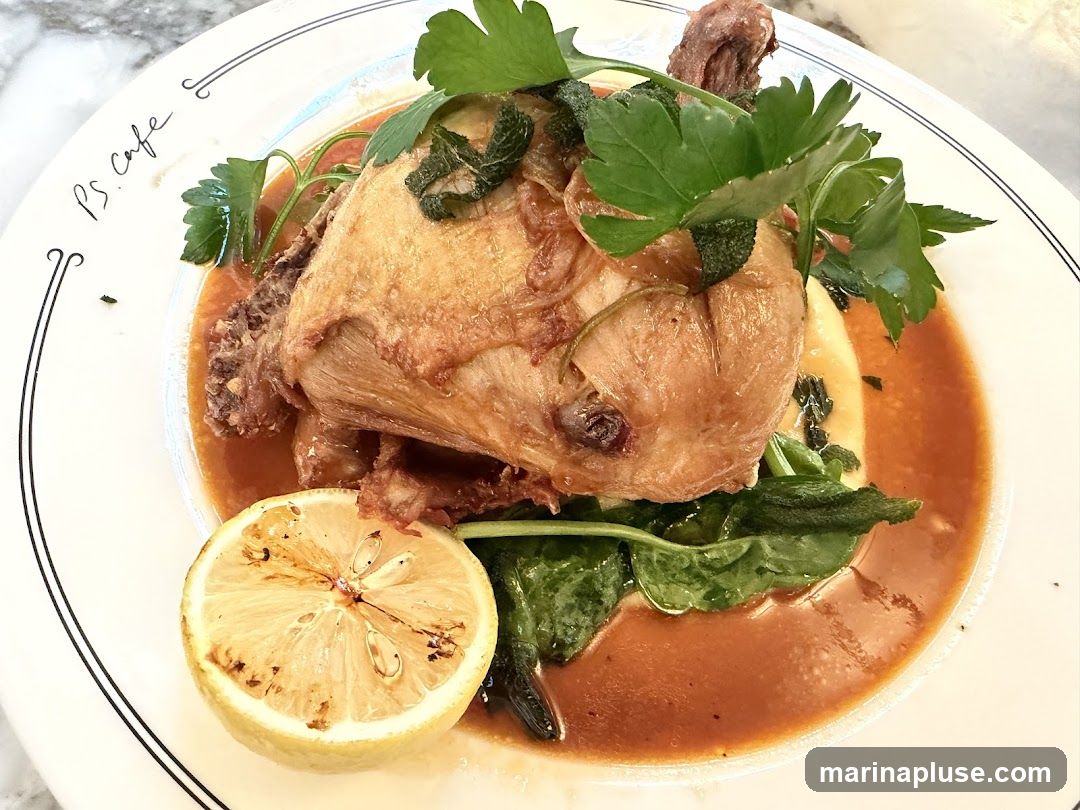 A close-up overhead view of a plate of roast chicken served with a rich sauce, polenta, wilted greens, and a grilled lemon half. The plate has a decorative rim with the text "ps. cafe" written in cursive.