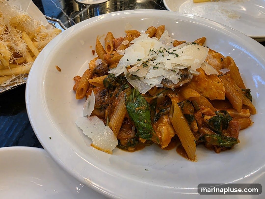 A close-up shot of a white bowl filled with penne pasta. The pasta is coated in a creamy red sauce and mixed with pieces of chicken, spinach, and mushrooms. It is topped with shaved parmesan cheese and a sprinkle of herbs. To the left, a silver tray holds a portion of french fries, also topped with shredded cheese.