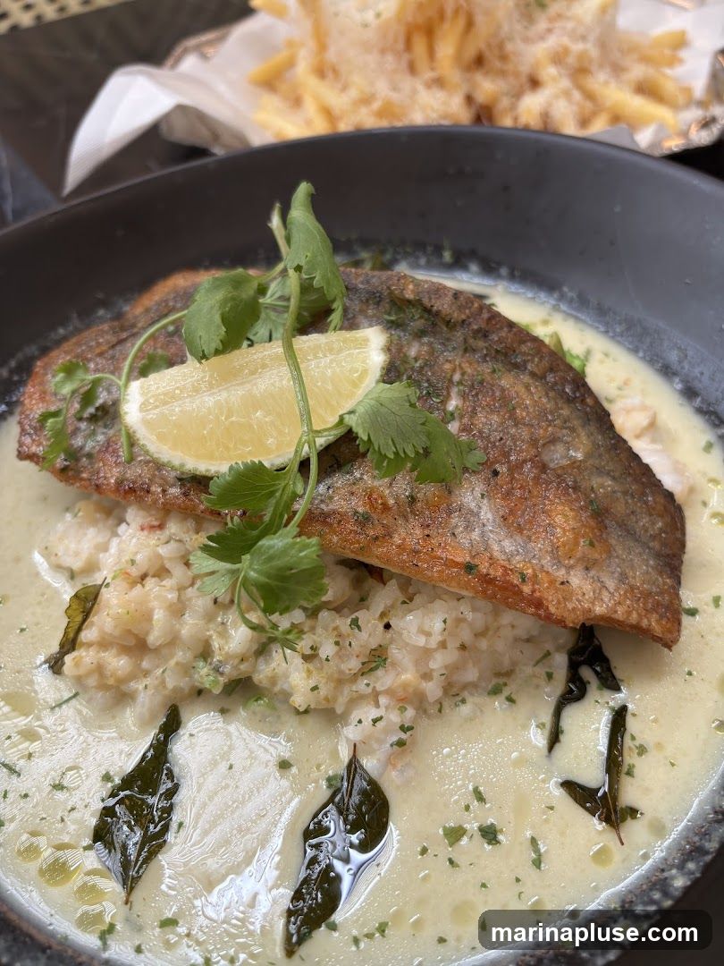 A close-up shot of a dish featuring a piece of pan-seared fish served over rice with a creamy sauce. The fish has crispy skin and is garnished with a lime wedge and cilantro. In the background, a serving of french fries topped with cheese is visible.