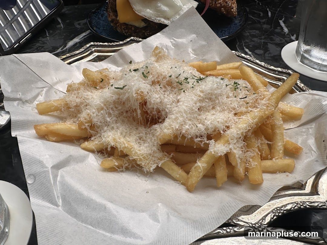A close-up shot of a serving of truffle parmesan fries, generously topped with grated cheese and herbs, presented on a white paper liner within a decorative silver tray. In the background, a burger with a fried egg and cheese is visible, along with a glass of water.