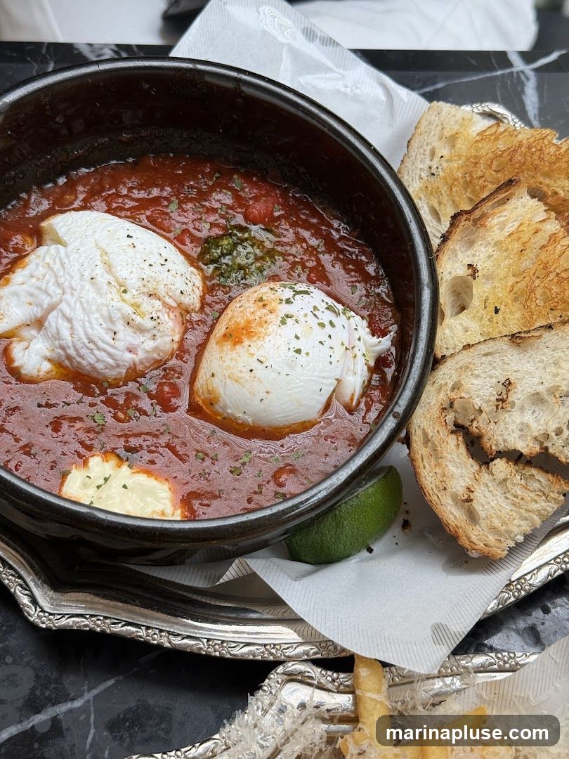 A close-up shot of a dish featuring two poached eggs nestled in a rich tomato sauce, garnished with herbs and a dollop of white sauce. Toasted bread slices are arranged on the side, and a wedge of lime is visible. The dish is served on a decorative silver tray with a white paper liner. In the foreground, a portion of french fries dusted with grated cheese is also visible.