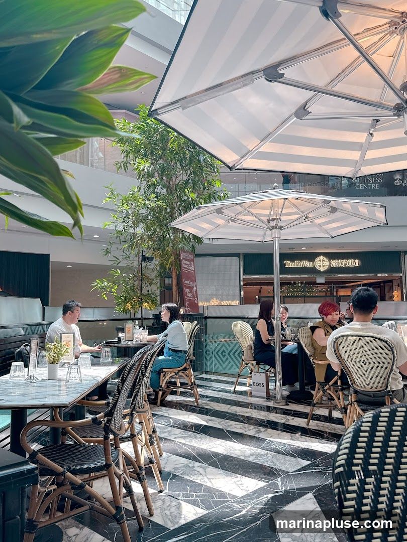 People are seated at tables in an outdoor cafe area, with striped umbrellas providing shade. The floor is black and white marble with a striped pattern. In the background, there are shops and a tree.