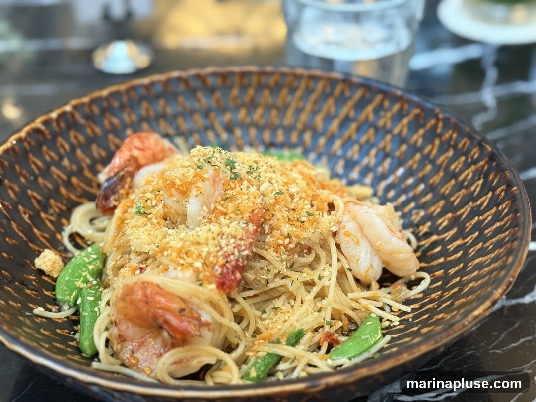 A close-up shot of a pasta dish with shrimp and snap peas in a decorative bowl. The pasta is topped with breadcrumbs and herbs.