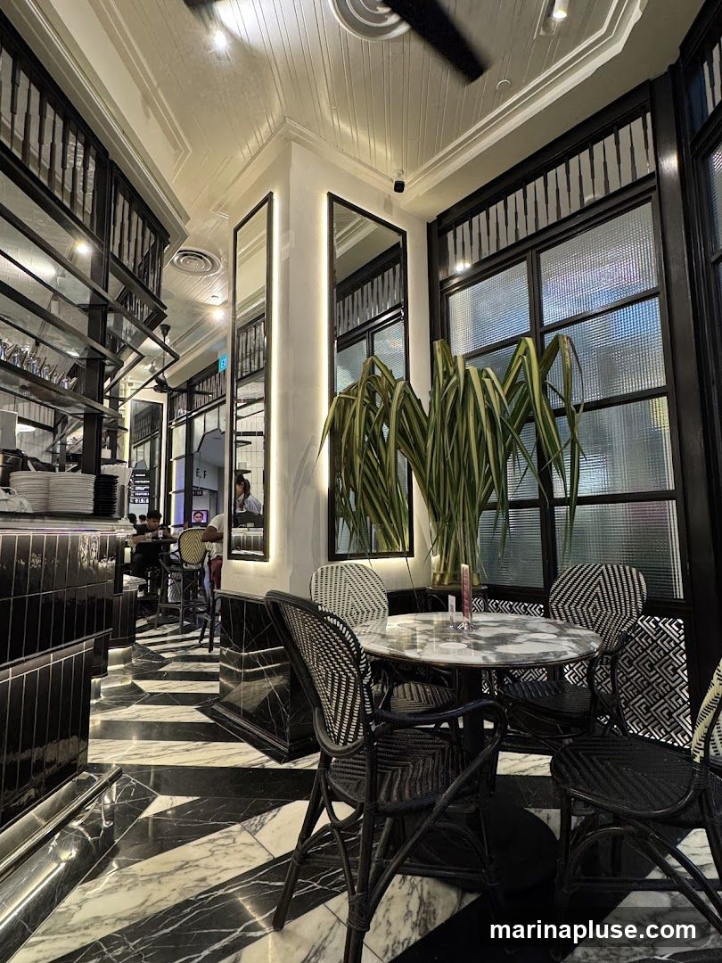 A view of a restaurant interior with black and white marble flooring, black wicker chairs around a marble-top table, and large windows with decorative grilles. Tall green plants add a touch of nature to the sophisticated decor. Mirrors on the wall reflect the elegant ambiance.