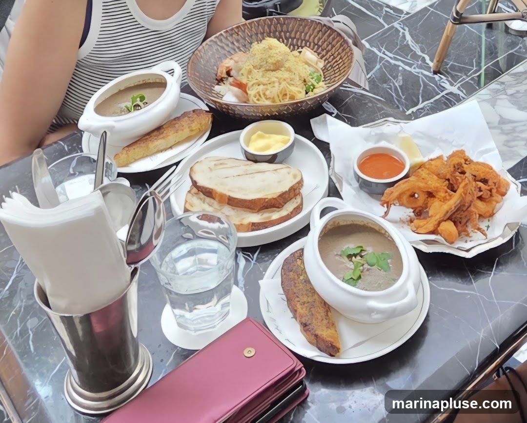A table is set with several dishes, including soup, sandwiches, pasta, and fried seafood. A person is partially visible on the left side of the image, wearing a striped tank top. The table has a black and white marble pattern.