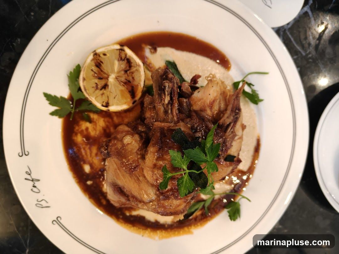 A close-up overhead shot of a roasted chicken dish served on a white plate with a dark sauce, a dollop of mashed potatoes or polenta, a grilled lemon slice, and garnished with parsley. The plate has a black rim with a decorative pattern and the text "ps. cafe" is visible on the left side.