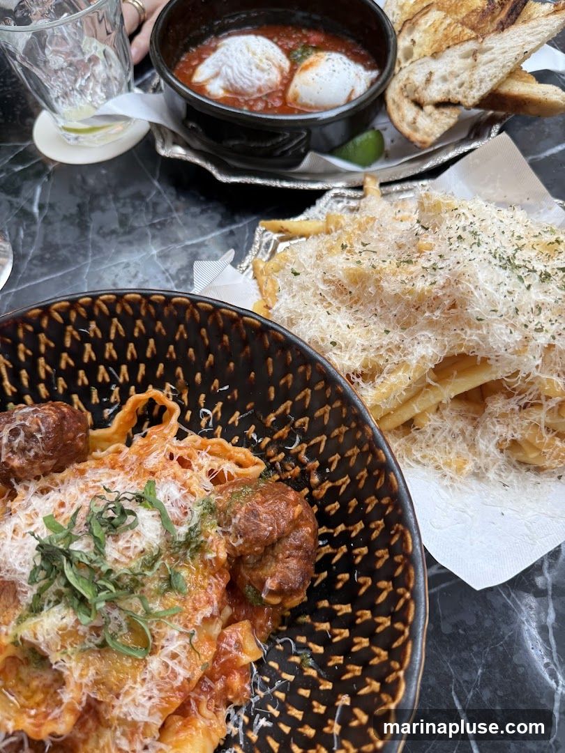 A close-up shot of a meal featuring pasta with meatballs and a side of fries topped with grated cheese. In the background, there is a dish with poached eggs in tomato sauce and toasted bread, along with a glass of water with lime.