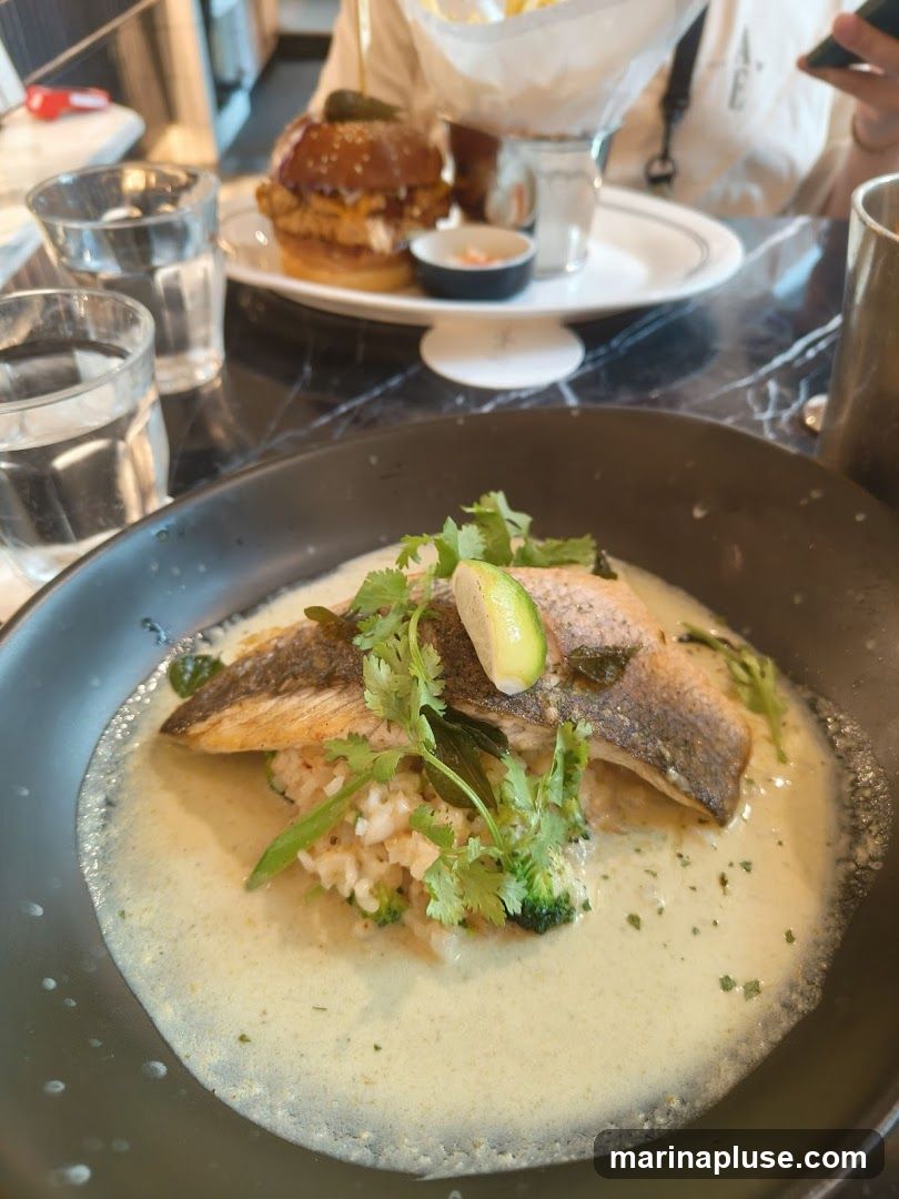 A close-up shot of a fish dish served in a black bowl. The fish fillet is placed on a bed of rice and broccoli, covered in a creamy green sauce. It is garnished with cilantro and a slice of lime. In the background, a burger and fries are visible on a white plate, along with two glasses of water.