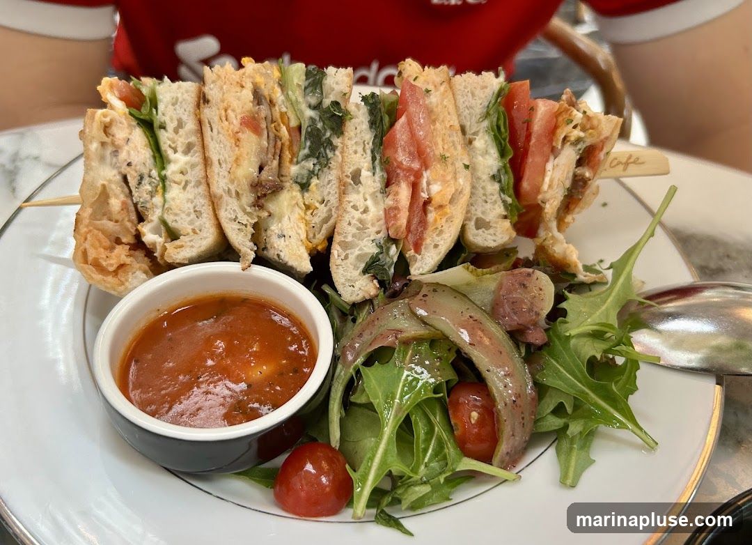 A club sandwich cut into quarters and served on a skewer, accompanied by a small bowl of tomato sauce and a side salad with cherry tomatoes and greens. The sandwich appears to be made with toasted bread and filled with lettuce, tomato, and meat. A person wearing a red shirt is visible in the background. A small wooden skewer with the word "cafe" is stuck in the sandwich.