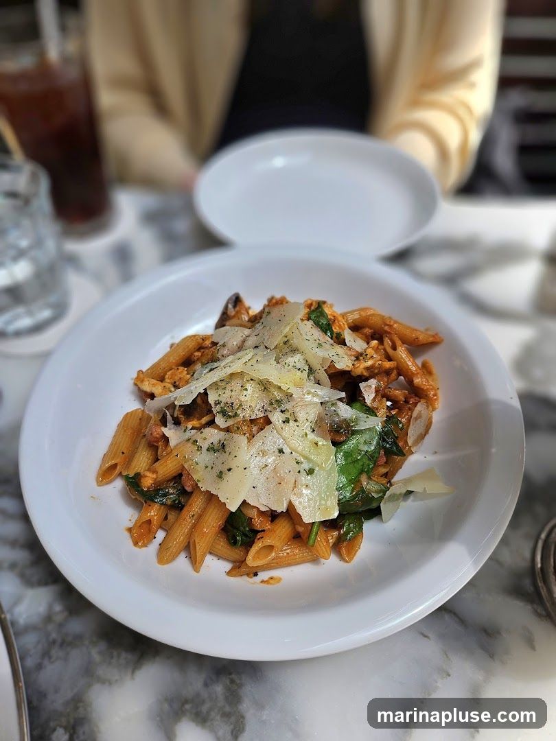 A close-up shot of a white bowl filled with penne pasta. The pasta is coated in a red sauce and mixed with spinach and topped with shaved parmesan cheese and herbs. In the background, a person is out of focus, wearing a light-colored cardigan over a dark top. A glass of iced beverage and a white plate are also visible on the marble-patterned table.