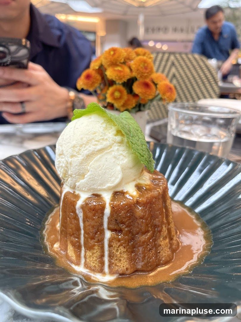 A close-up of a dessert, likely a sticky toffee pudding, served in a dark, fluted bowl. The dessert is topped with a scoop of vanilla ice cream and a sprig of mint. A caramel sauce is drizzled over the pudding and pooled around its base. In the background, a person is visible holding a phone, and a vase of orange flowers sits on the table. Another person is seated further back in what appears to be an outdoor dining area.
