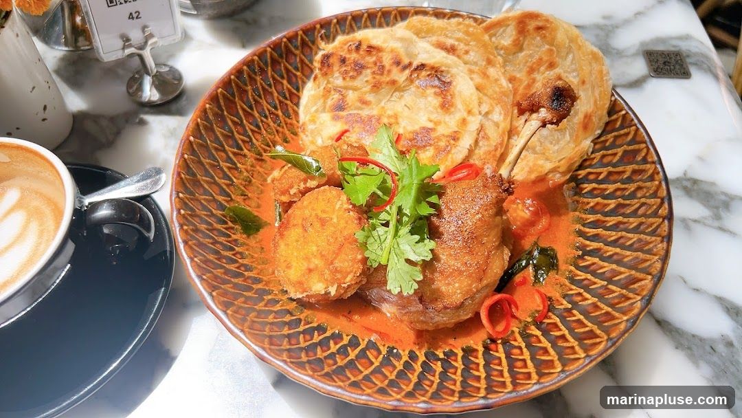 A close-up shot of a dish featuring duck curry served with roti bread. The curry is a vibrant orange color and contains pieces of duck, fried dumplings, cherry tomatoes, and garnished with cilantro and chili slices. A cup of coffee with latte art is visible in the foreground. The meal is presented on a patterned plate and set on a marble-patterned table. A small QR code stand is also visible on the table.