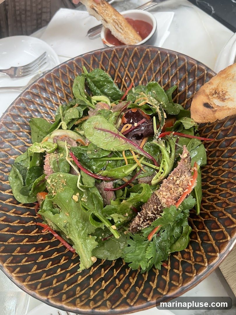 A close-up overhead shot of a beef salad served in a textured brown bowl. The salad contains mixed greens, thinly sliced beef, shredded carrots, and red cabbage. It is garnished with chopped nuts and served with a side of toast and a small dish of red sauce.