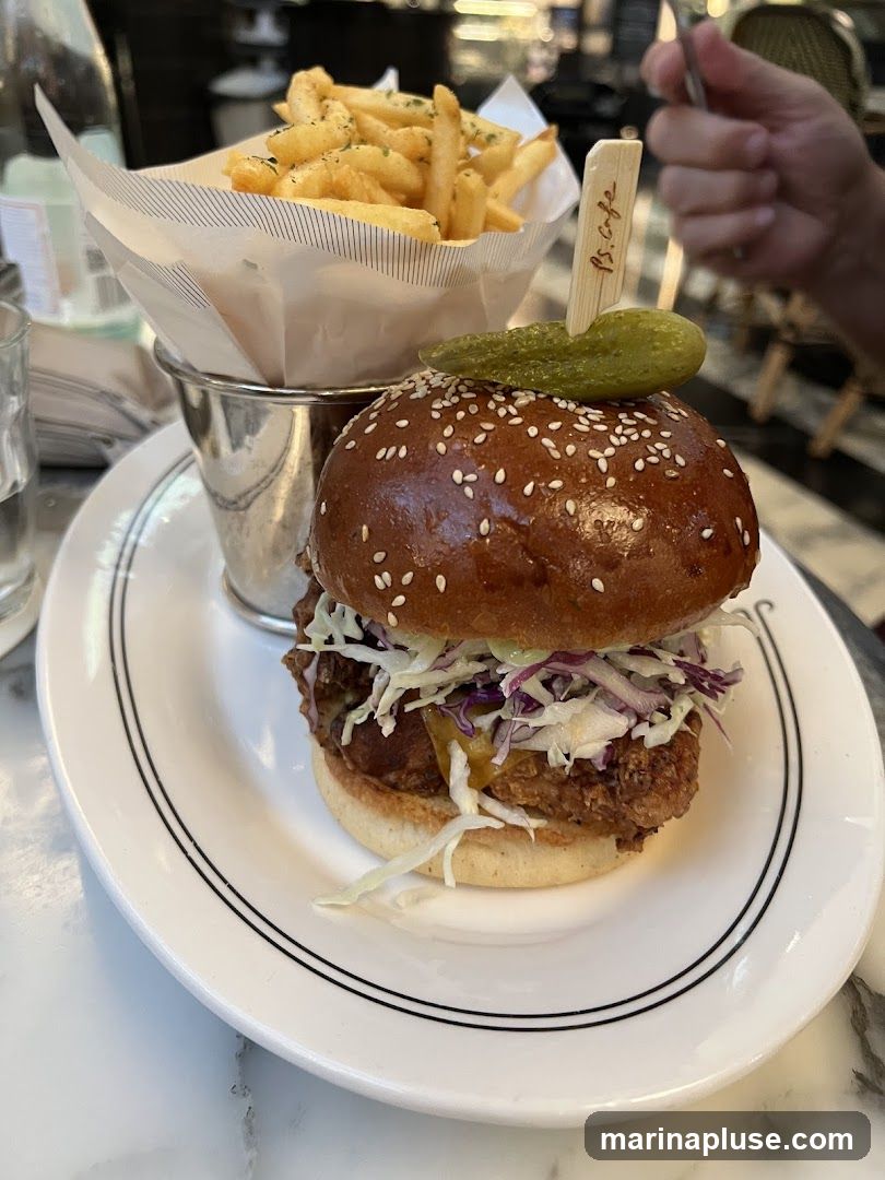 A close-up shot of a fried chicken burger served on a white plate with a side of french fries in a metal bucket lined with paper. The burger has a sesame seed bun, coleslaw, and a pickle spear on top. A small wooden skewer with the text "PS. Cafe" is stuck into the pickle. The background shows a blurred restaurant interior.