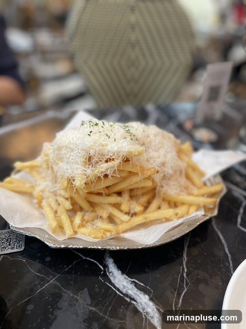A close-up shot of a plate of truffle fries, generously topped with grated cheese and parsley. The fries are golden brown and appear crispy. The plate is placed on a black marble table. In the background, a blurred image of a restaurant interior is visible, with a decorative lamp and patrons.