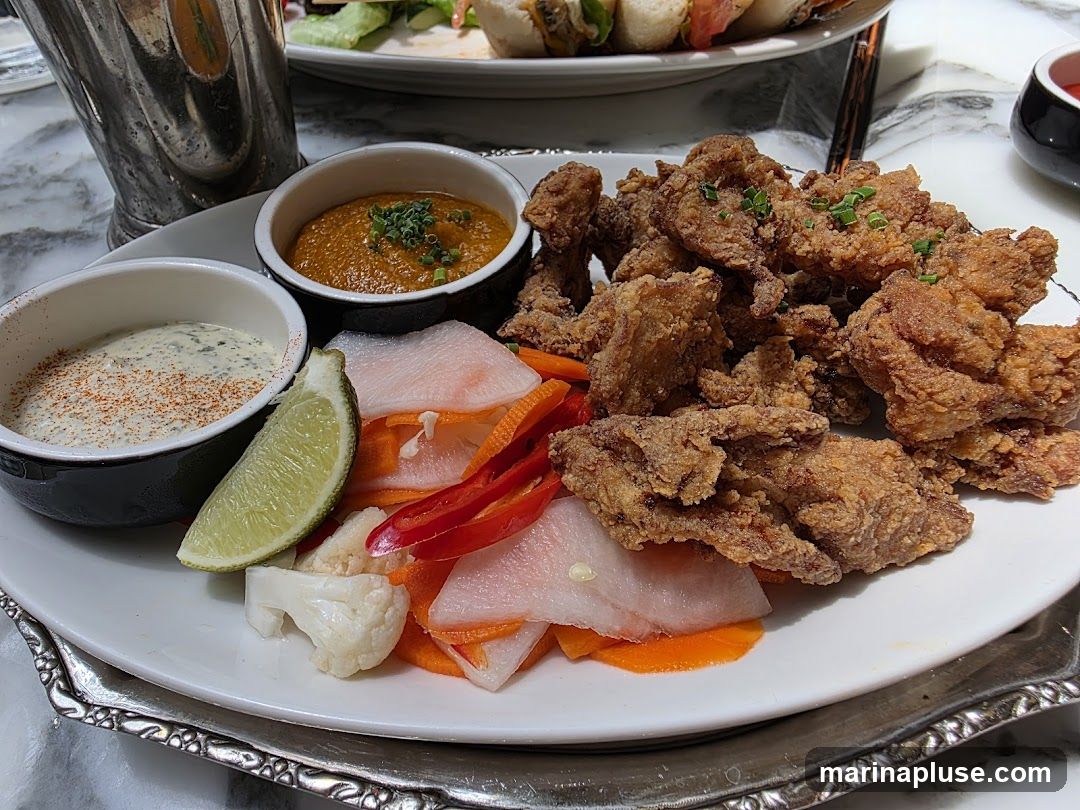 A plate of crispy fried chicken pieces served with two dipping sauces, pickled vegetables including carrots, radishes, cauliflower, and chili peppers, and a lime wedge. A metal shaker and another plate with food are visible in the background.