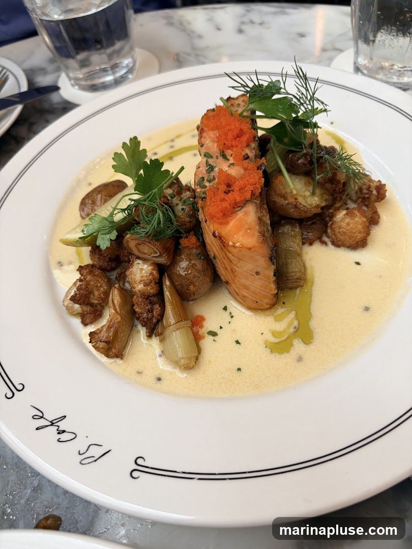 A plate of salmon served with roasted potatoes, cauliflower, and leeks in a creamy sauce. The dish is garnished with fresh herbs and roe. The plate has the inscription "P.S. Cafe" on its rim.