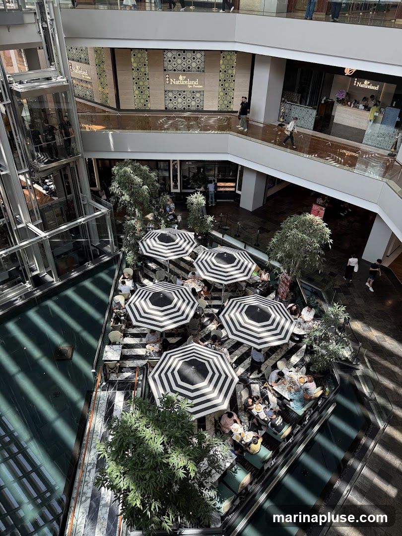 An aerial view of a cafe situated in an indoor courtyard of a multi-level building. The cafe features black and white striped umbrellas providing shade over tables where patrons are seated. Lush green trees are interspersed around the seating area. In the background, a spa with signage is visible on an upper level, along with an elevator and other building interiors.