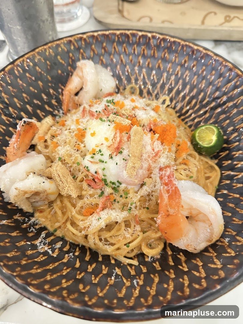 A close-up shot of a bowl of spaghetti with shrimp, topped with grated cheese, roe, and a poached egg. A lime wedge is on the side.