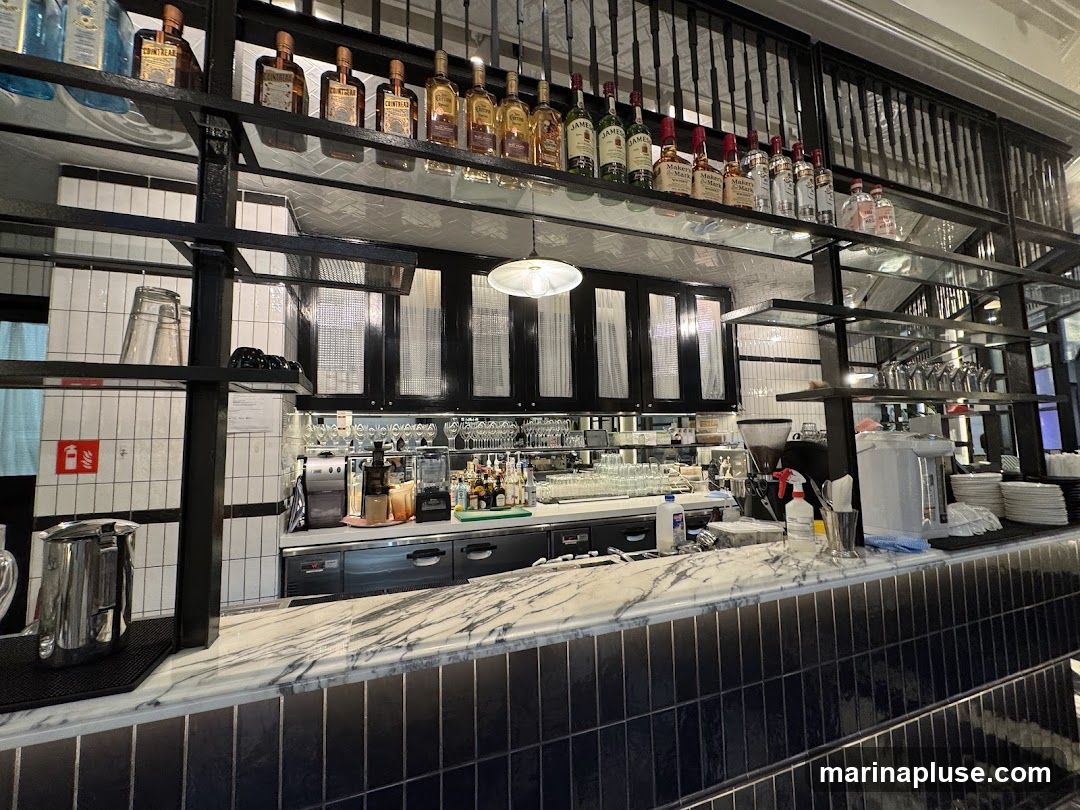 A well-lit bar counter with various bottles of alcohol displayed on shelves above. The counter itself is made of marble with a tiled backsplash. Behind the counter, there are coffee machines, blenders, and stacks of glasses and plates.