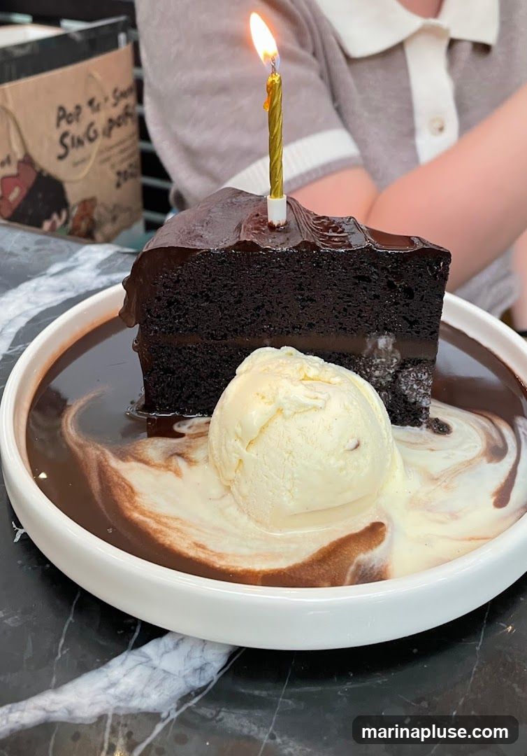 A slice of rich chocolate lava cake is served in a white bowl with a scoop of vanilla ice cream. The cake is topped with a lit birthday candle and sits in a pool of warm chocolate sauce, swirled with cream. In the background, a paper bag with text is visible.