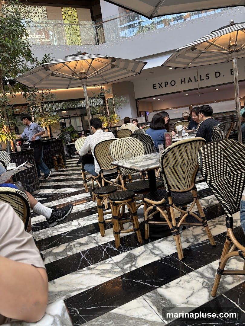 An outdoor seating area of a cafe with black and white striped marble flooring. Tables and chairs with woven backs are set up under striped umbrellas. Several patrons are seated at the tables, and a server is visible in the background. In the distance, signage reads "EXPO HALLS D, E".