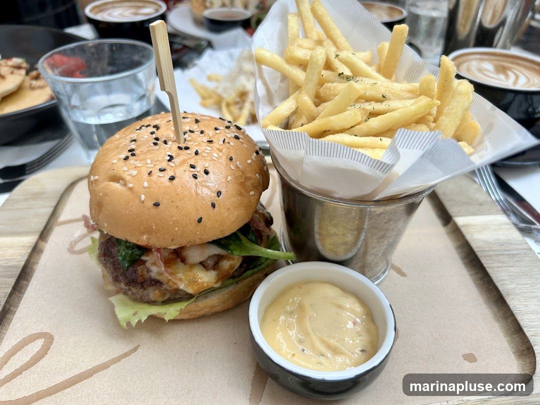 A close-up shot of a gourmet burger served with a side of french fries and dipping sauce. The burger is on a sesame seed bun with lettuce, cheese, and a patty. The fries are in a metal bucket lined with white paper. A small bowl of sauce sits in front of the burger. In the background, there are cups of coffee and a glass of water.