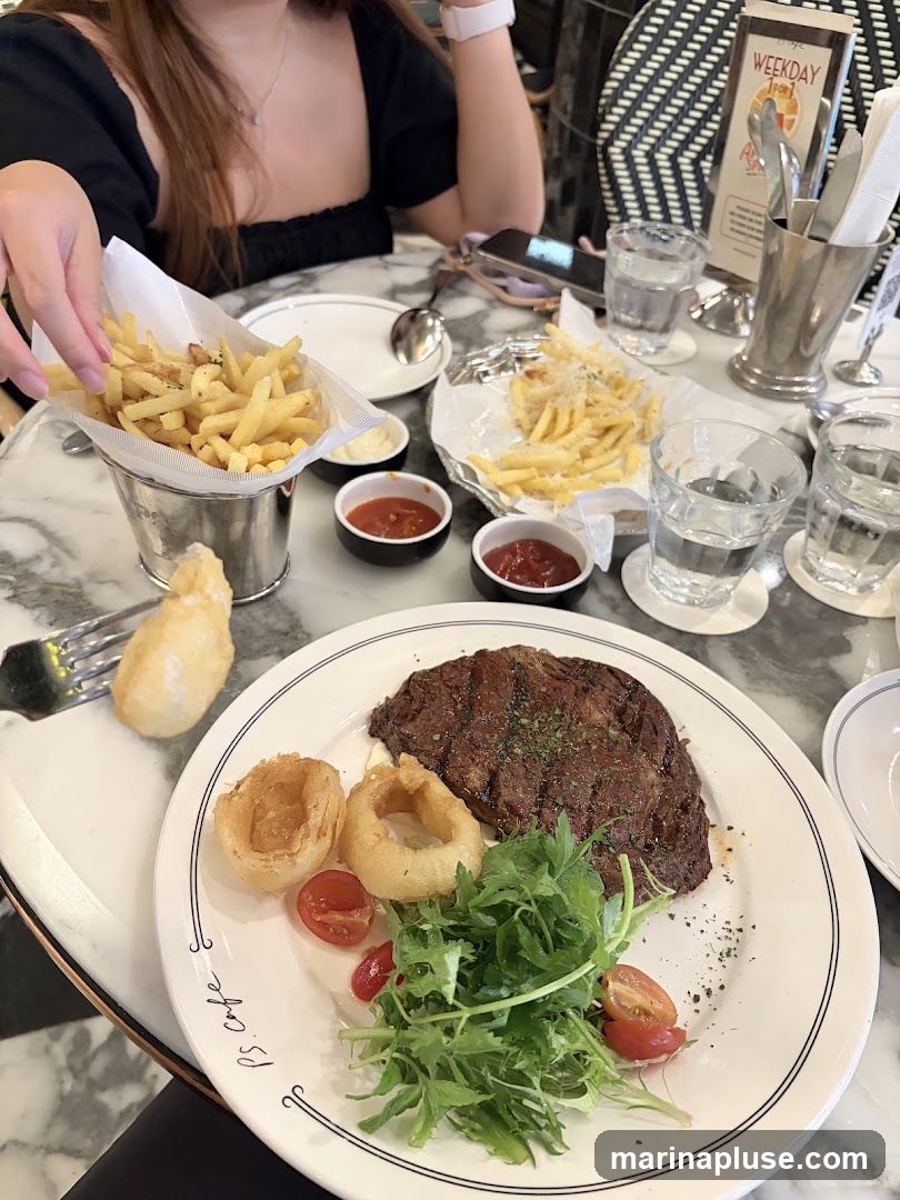 A close-up shot of a steak dinner served on a white plate with a side of fries and onion rings. The steak is grilled and seasoned with herbs, accompanied by a fresh green salad and cherry tomatoes. Two servings of fries are visible, one in a metal bucket and another on a foil-lined plate, with dipping sauces. The setting appears to be a restaurant with marble tabletops and a menu stand in the background.