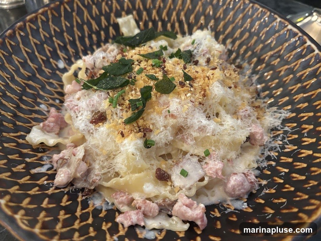 A close-up shot of a pasta dish served in a dark, textured bowl. The pasta appears to be a wide, flat noodle, possibly lasagna, coated in a creamy sauce. It is topped with grated cheese, crispy breadcrumbs, fresh sage leaves, and chopped chives. Small pieces of meat, likely pork or bacon, are mixed throughout the dish.