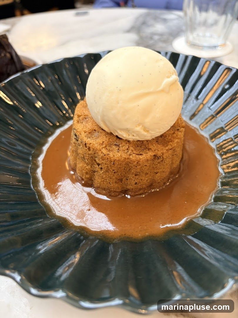 A close-up shot of a dessert served in a dark, fluted plate. The dessert is a small, round cake or pudding, topped with a scoop of vanilla ice cream. The cake is surrounded by a pool of caramel or butterscotch sauce. The background is blurred, showing a table setting with a glass and a hint of other dishes.