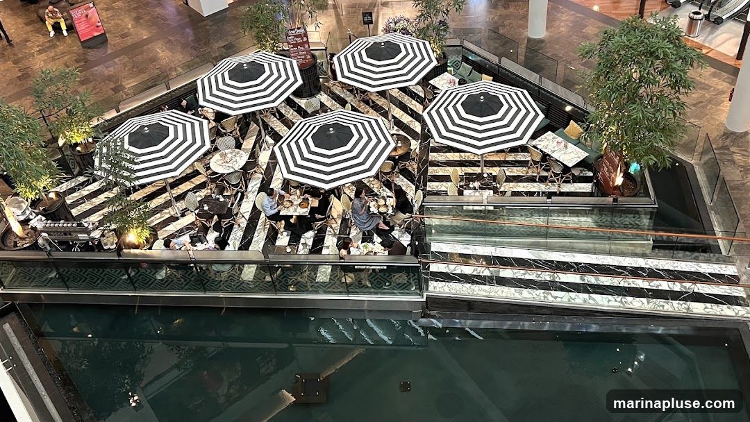 An aerial view of an outdoor cafe seating area with black and white striped umbrellas and tables. The cafe is situated in a modern indoor space with a reflective pool below. Several patrons are seated at the tables, enjoying their meals and drinks. The decor features a striking black and white striped floor pattern and potted plants. A coffee machine is visible on the left side of the seating area.