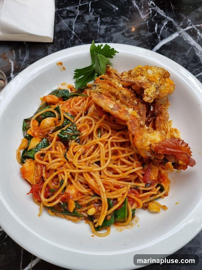 A white bowl filled with spaghetti tossed in a red sauce with spinach and shrimp. A whole fried soft shell crab is placed on top of the pasta. A sprig of parsley garnishes the dish. The table surface is black marble.