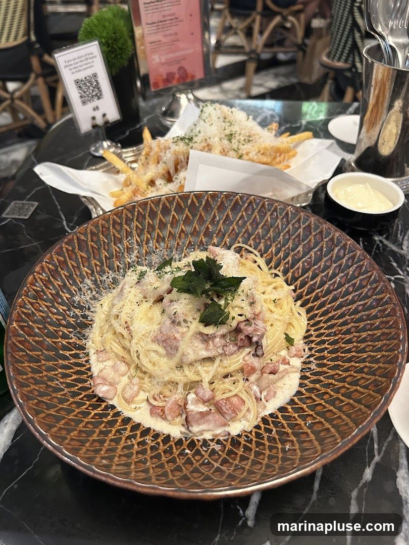 A close-up view of a plate of spaghetti carbonara and a side of french fries, both topped with grated cheese and herbs. A small dish of dipping sauce is also visible. The food is served on a marble-patterned table.