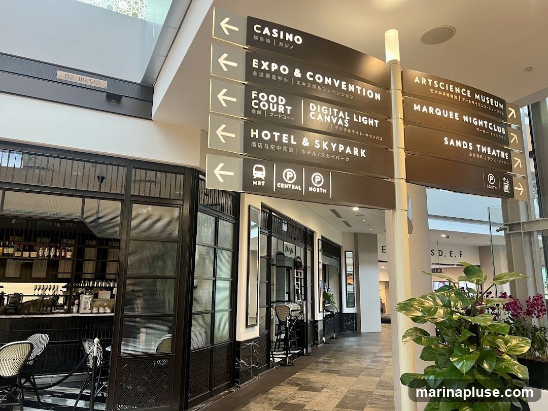 A multi-directional signpost in a modern indoor space, indicating various attractions and facilities such as CASINO, EXPO & CONVENTION, FOOD COURT, DIGITAL LIGHT CANVAS, HOTEL & SKYPARK, MRT, ARTSCIENCE MUSEUM, MARQUEE NIGHTCLUB, and SANDS THEATRE. The signage includes text in English, Chinese, and Japanese. In the foreground, a cafe or restaurant with black and white checkered flooring and seating is visible.