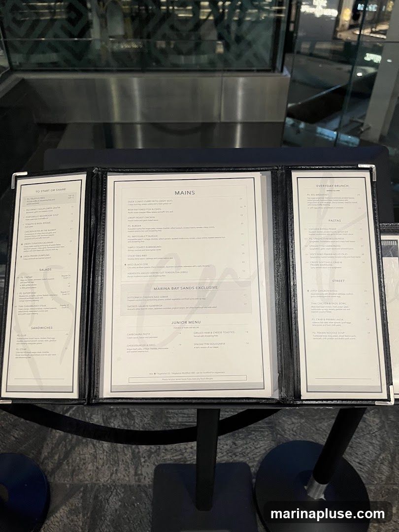 A multi-panel menu displayed on a stand. The menu is divided into sections including 'To Start or Share', 'Salads', 'Sandwiches', 'Mains', 'Marina Bay Sands Exclusive', 'Junior Menu', 'Everyday Brunch', 'Pastas', and 'Street'.