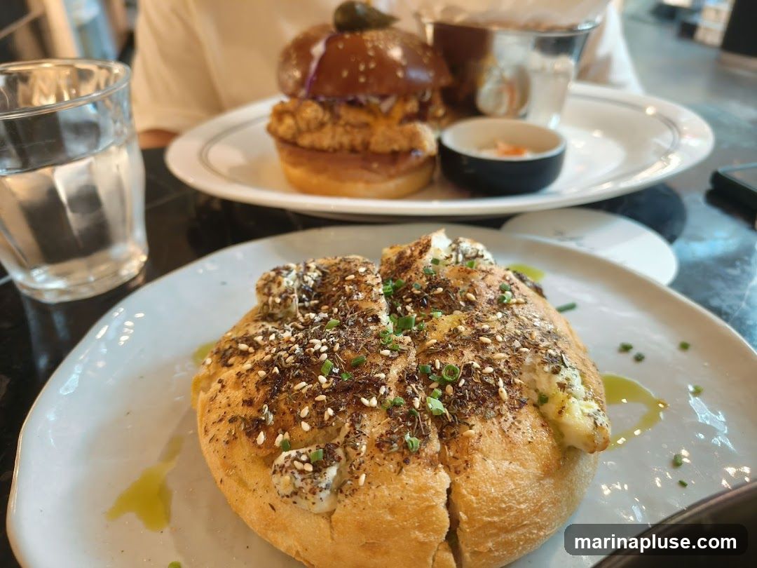 A close-up shot of two plates of food, with a glass of water in the foreground. The plate in the foreground features a round bread item topped with herbs, seeds, and a creamy spread. The plate in the background holds a burger with a pickle on top, and a small dish of sauce.