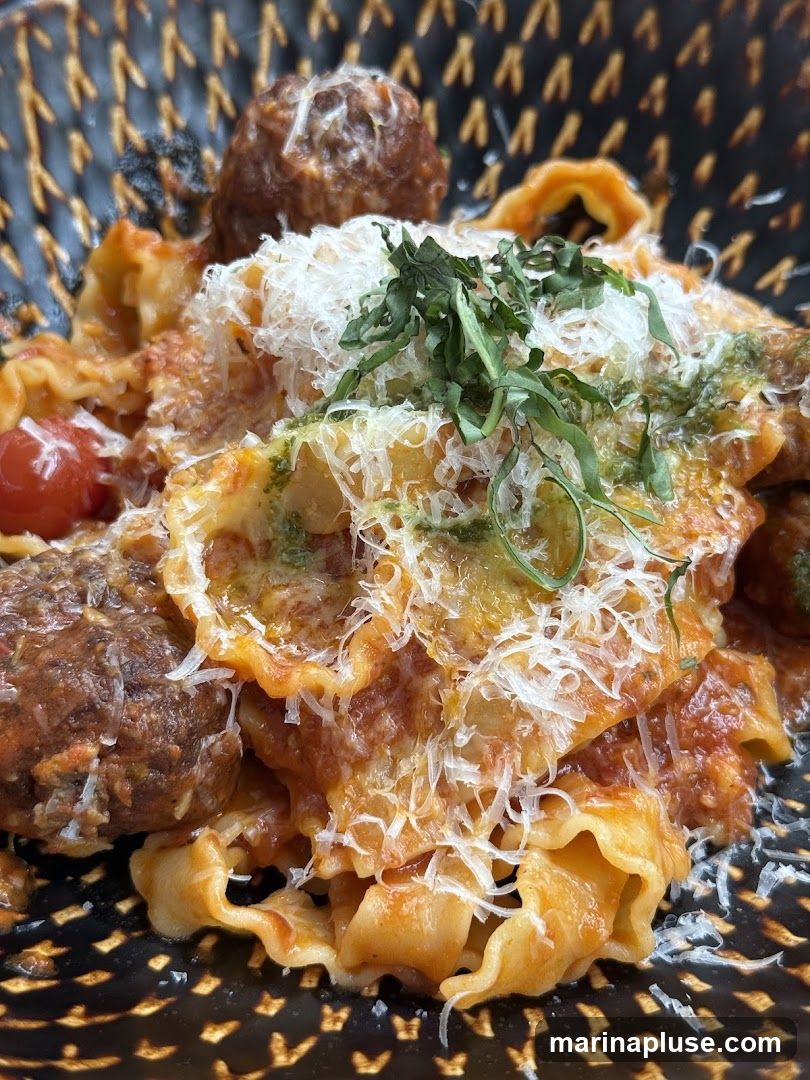 A close-up shot of a bowl of pasta with meatballs. The pasta appears to be a ruffled or wavy shape, coated in a rich tomato sauce. Several large meatballs are visible, along with a sprinkle of grated cheese and fresh green herbs. A few cherry tomatoes are also present in the dish. The bowl has a textured, dark pattern.