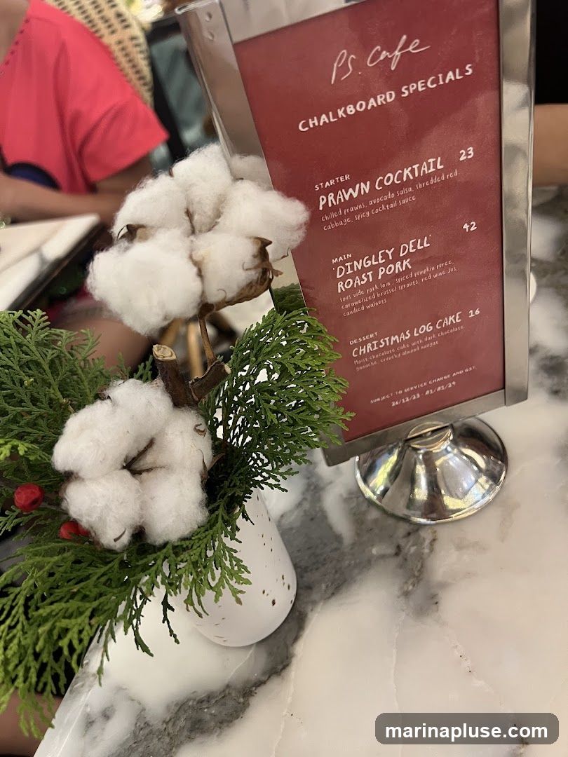 A menu board displaying specials from P.S. Cafe, featuring a starter, main course, and dessert. The menu is presented in a silver frame on a marble table, with a floral arrangement of cotton and greenery in the foreground.