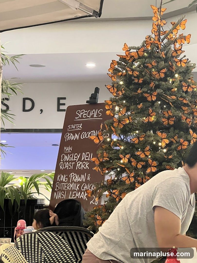 A chalkboard menu board displays specials, including starters and main courses. A decorated Christmas tree with monarch butterflies and lights is visible to the right. People are seated at tables in the foreground.