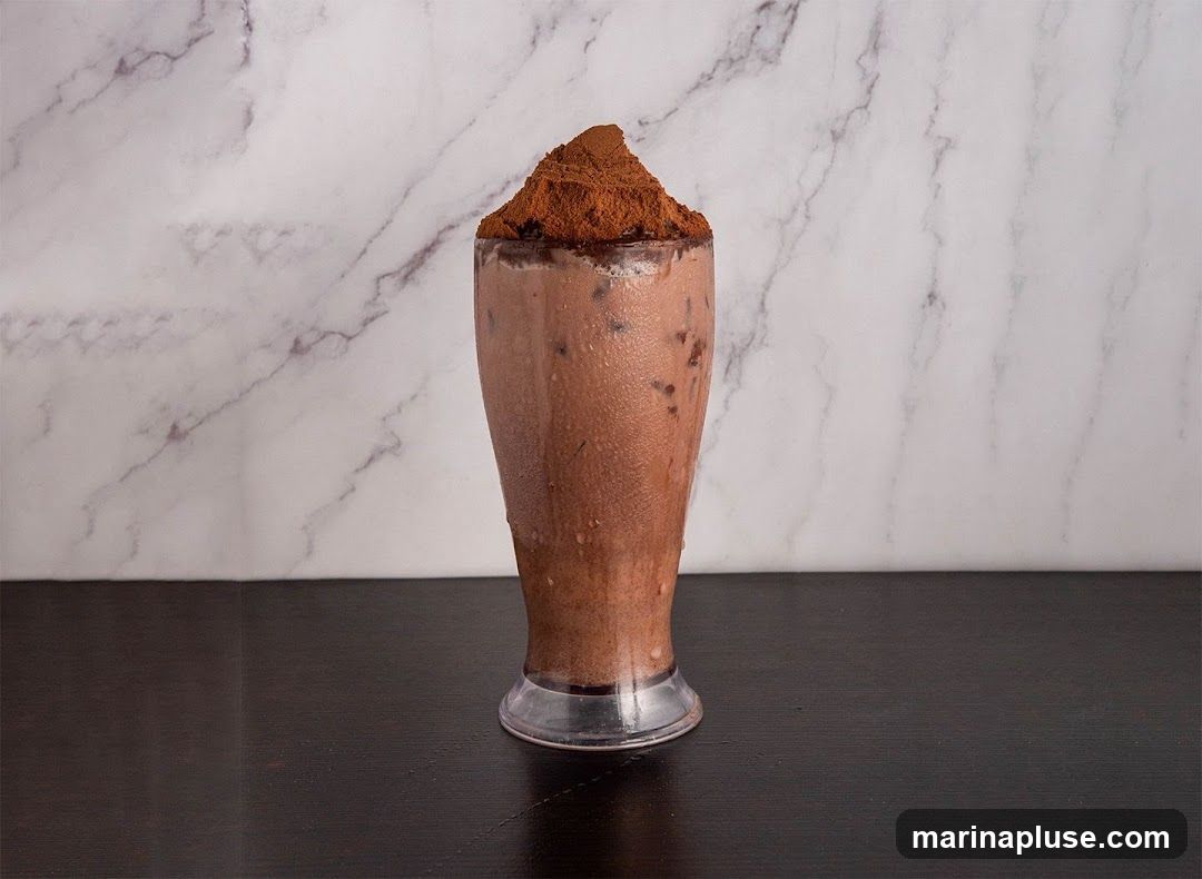 A tall glass of chocolate drink, possibly a milkshake or smoothie, is filled with ice and chocolate chunks. The drink is topped with a generous mound of cocoa powder. The background features a marble-patterned wall and a dark wooden surface in the foreground.