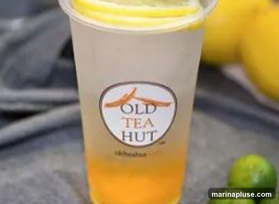 A close-up shot of a clear plastic cup filled with a layered beverage, likely lemon tea. The drink has a yellow-orange base and a lighter, translucent top, garnished with lemon slices. A circular logo on the cup reads "OLD TEA HUT" with a mountain-like graphic above the text. Several limes and lemons are visible in the blurred background.