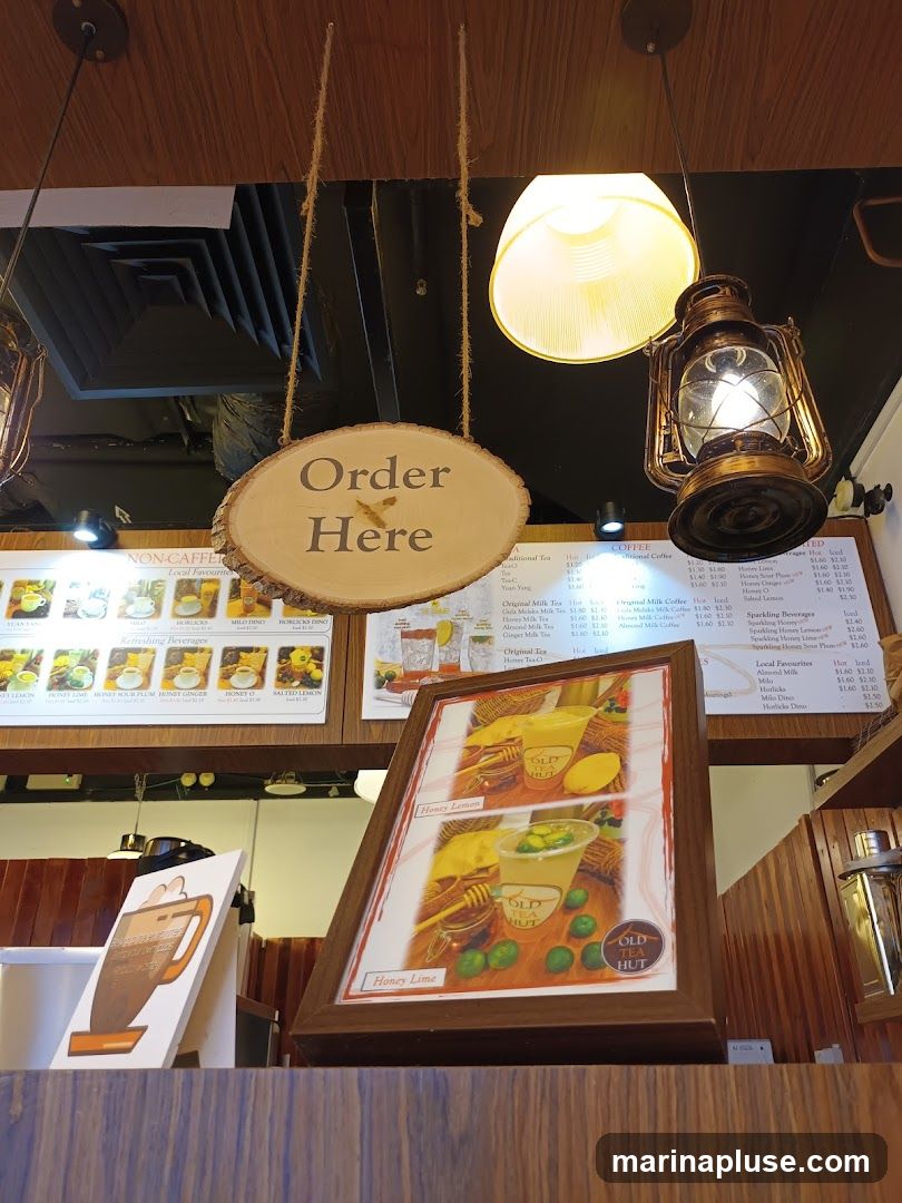 A wooden sign with 'Order Here' hangs above a display of menus and framed pictures of drinks. The menus show various beverages including traditional teas, coffees, milk teas, and sparkling drinks. Featured drinks include Honey Lemon and Honey Lime.