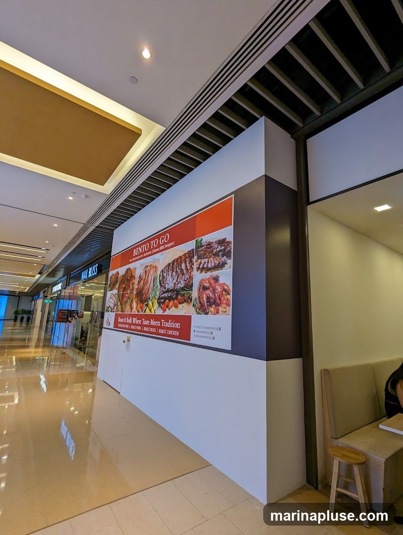 A large advertisement for "Bento To Go" featuring images of various roasted meats, including roast pork, roast duck, and roast chicken. The text highlights "Where Taste Meets Tradition" and lists the types of roast meats offered. The advertisement is displayed on a wall in a shopping mall corridor.
