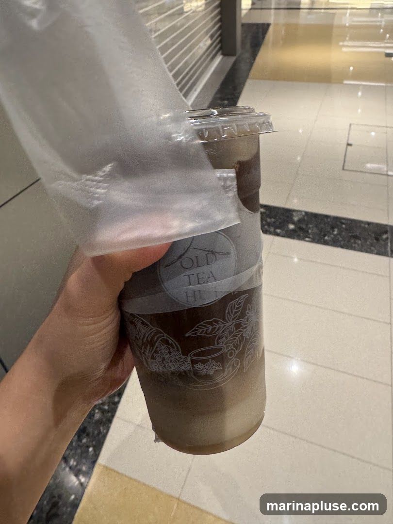 A hand holds a clear plastic cup filled with an iced beverage. The cup has a white sleeve with a logo that reads "OLD TEA HUT" and features illustrations of a cornucopia, tea leaves, and a teacup. The background appears to be an indoor walkway with tiled flooring and closed storefronts.