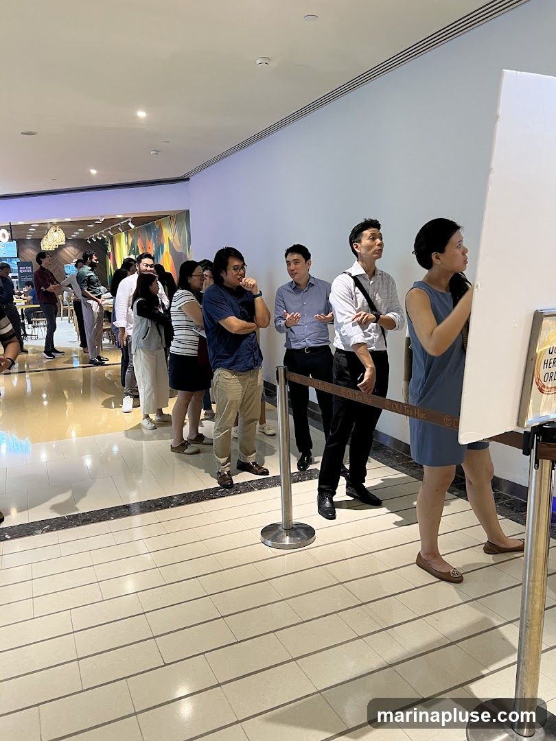 A group of people are standing in a queue, waiting to be served. The queue is indoors, possibly in a mall or food court. Some individuals are looking at a white board or menu. The floor is tiled with a reflective surface.